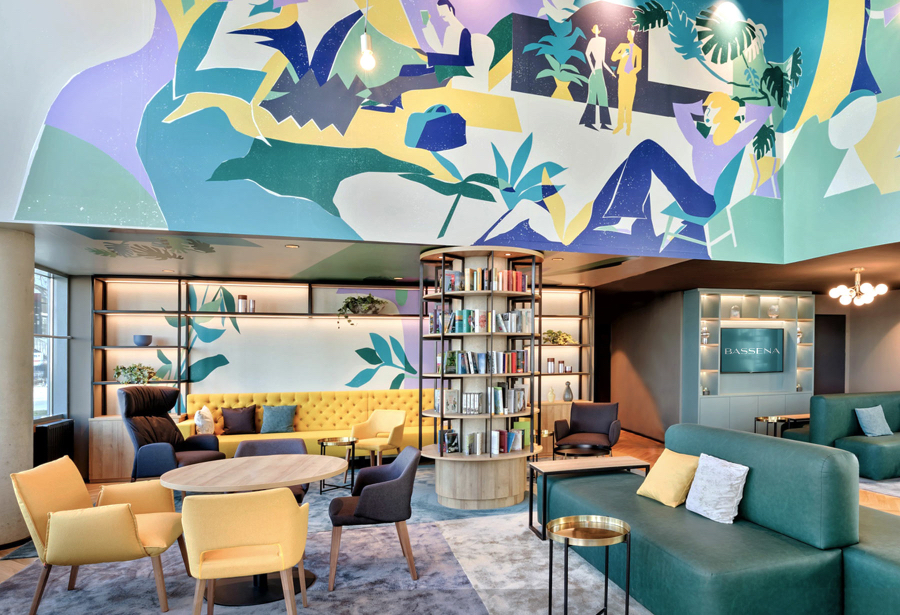 Lobby of the Vassena Hotel. This space is decorated with a variety of different colors, giving it a refreshed look, we have a yellow sofa and some yellow chairs, but also a green sofa and grey chairs. There's mural on the wall above it with the color of the rooms. kitzig design studios Kitzig Design Studios- From Germany to the World Interior Bassena Wien 03 1 1