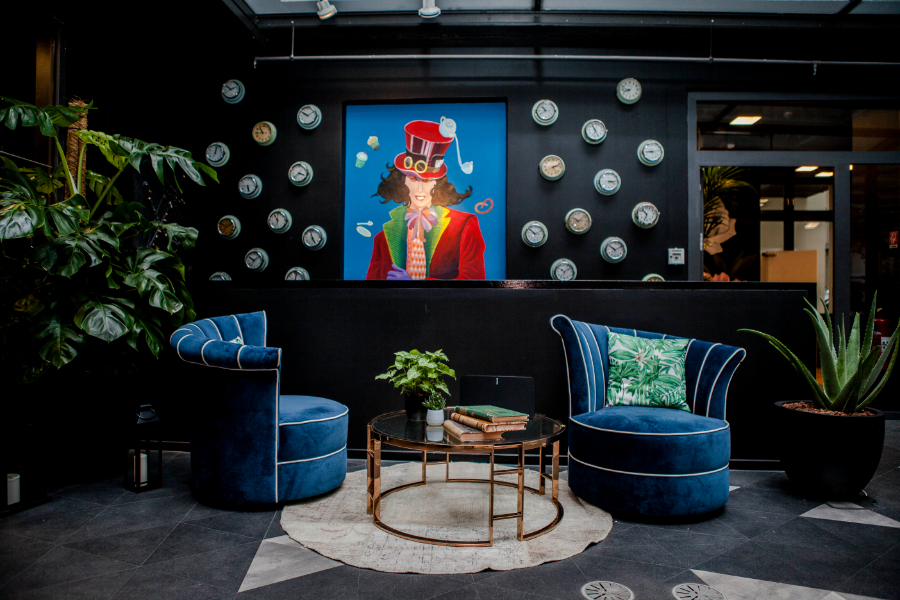 Ivy's Design - The twist we didn't know we needed Alice in Wonderland. Here we see a painting of the Mad Hatter on the wall, two big blue armchairs, some plants, a round table with books in the middle and clocks on the wall.  ivy's design Ivy’s Design – The twist we didn’t know we needed The Twist of Ivys Design in Modern Interior Decor Alice in Wonderland decoration