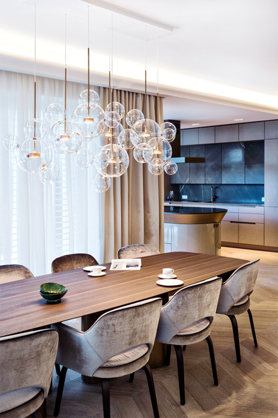 PRIVATHAUS SCHÖNE AUSSICHT dining room with a transparent chandelier, light dining chairs, a light wooded table, and a view of the kitchen. modern interior design ideas Reimann Architecture: Modern Interior Design Ideas Privathaus SchoeneAussicht  Reimann Architecture Modern Interior Design Ideas